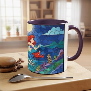 Puede incluir: Una taza de cer&aacute;mica blanca con interior y asa morados, con un colorido dise&ntilde;o de vidriera de Ariel de La Sirenita. El dise&ntilde;o incluye un cangrejo, coral y el nombre "thia".