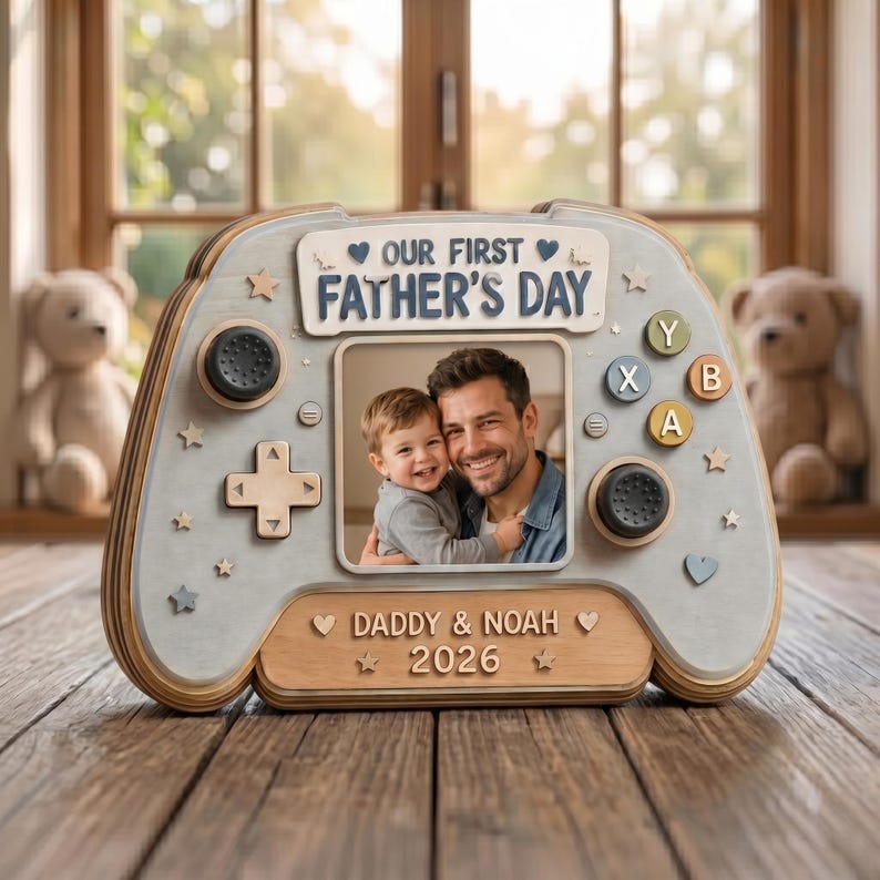 May include: A wooden photo frame shaped like a video game controller. The frame is light gray with wooden accents and features the text "OUR FIRST FATHER'S DAY" and "DADDY & NOAH 2026". A photo of a father and child is in the center.