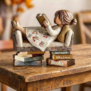 Può includere: Statuina di una ragazza che legge un libro mentre è sdraiata su un piccolo divano. Il divano è sostenuto da pile di libri. La ragazza indossa un vestito bianco con un motivo floreale. Il testo sui libri recita "Just a Girl Who Loves Books."