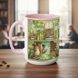 Pu&ograve; includere: Tazza in ceramica bianca con interno e manico rosa. La tazza presenta un'illustrazione colorata di una scena di biblioteca con libri, una rana e una donna in abito verde che legge. La tazza &egrave; appoggiata su un tavolo di legno.