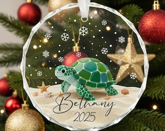 May include: A clear, circular Christmas bauble with a cut-glass edge, depicting a green turtle on a sandy beach with snowflakes. The name "Bethany" and the year "2025" are written on the sand. A white ribbon is attached for hanging.