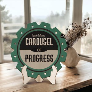 May include: A green and white wooden sign with a gear shape and the text "Walt Disney Carousel of Progress". The sign is on a wooden stand.