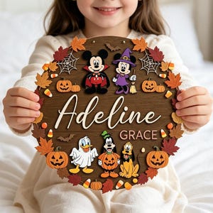 Pode incluir: Uma placa redonda de madeira com fundo de grão de madeira castanho escuro com o nome Adeline Grace em letras brancas e cor-de-rosa. A placa está decorada com folhas de outono coloridas, abóboras, doces, teias de aranha e personagens Disney.