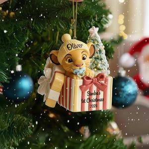 May include: A Christmas tree ornament depicting a Simba cub wearing a yellow hat with the name "Oliver". The cub is in a striped gift box with a red bow and the text "Simba's 1st Christmas 2025". A small Christmas tree and a blue dummy are also visible.