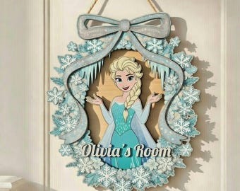 Op de afbeelding: Een houten deurbord met een cartoonafbeelding van Elsa uit Frozen, omringd door een krans van sneeuwvlokken en een lichtblauwe strik. Het bord leest "Olivia's Room" in een cursief lettertype en is opgehangen aan een touw.