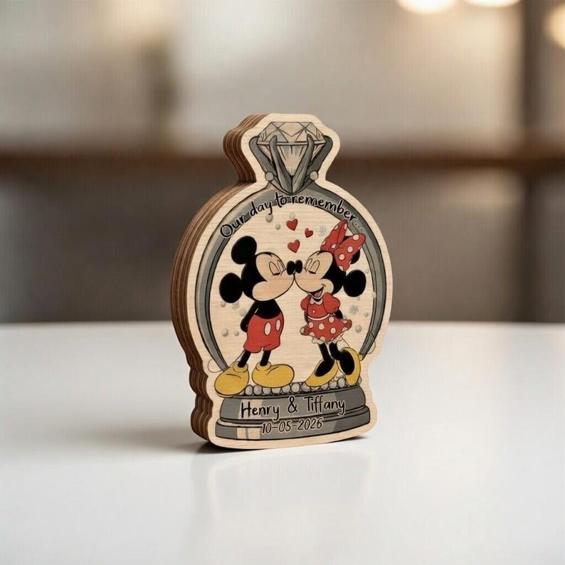 May include: Wooden engagement ring-shaped decoration featuring Mickey and Minnie Mouse kissing. The design includes the text "Our day to remember" and personalized names and date. The characters are in their classic outfits.