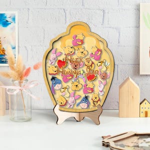 Puede incluir: Decoración de madera con forma de tarro de miel con personajes de Winnie the Pooh. El tarro tiene un marco marrón claro y un interior amarillo con ilustraciones de personajes y el texto "Alex's Hunny Jar". La decoración está soportada por un soporte de madera.