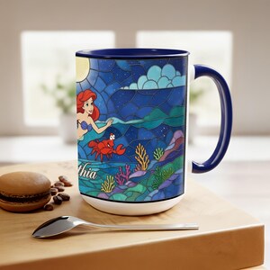 Puede incluir: Taza de cer&aacute;mica blanca con interior y asa azules. La taza presenta una ilustraci&oacute;n de estilo vidriera de una sirena, un cangrejo rojo y elementos submarinos. El nombre "Cynthia" est&aacute; impreso en la taza.