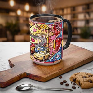 Puede incluir: Una taza de caf&eacute; de cer&aacute;mica con asa y borde negros, con un colorido dise&ntilde;o de vidriera. El dise&ntilde;o incluye im&aacute;genes de coches de carreras, corazones y una pista de carreras. La taza est&aacute; llena de caf&eacute; caliente y est&aacute; sobre una tabla de madera.