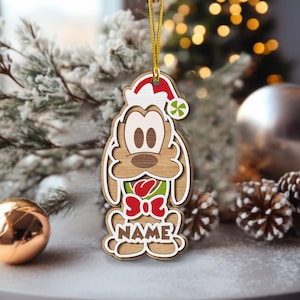 May include: Wooden Christmas ornament depicting a cartoon dog in a Santa hat and a red bow. The layered design ornament has the word "NAME" at the bottom. It is suspended by a gold string.