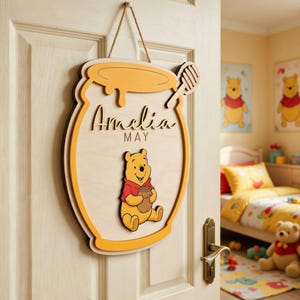 May include: Wooden honey pot-shaped sign with the name "Amelia" and "MAY" in script. Features a Winnie the Pooh character holding a honey pot. The sign is hanging on a white door with a rope.