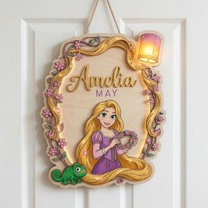 Op de afbeelding: Een houten wandbord met een stripfiguur met lang blond haar, een paarse jurk en een groene kameleon. Het bord toont de naam Amelia May, is versierd met roze bloemen en bevat aan de bovenkant een verlichte lantaarn als decoratie.