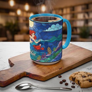 Puede incluir: Una taza de caf&eacute; de cer&aacute;mica con asa y borde azules, con un dise&ntilde;o de vidriera de Ariel de La Sirenita. El dise&ntilde;o incluye una sirena, un cangrejo y elementos submarinos. La taza est&aacute; llena de caf&eacute; y est&aacute; sobre una tabla de madera.