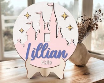 Pode incluir: Um sinal de madeira rosa e branco em forma de castelo com o nome "Lillian Kate" escrito em roxo. O sinal está em um pequeno suporte de madeira e tem estrelas douradas ao redor do castelo.