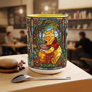 May include: A white ceramic mug with a yellow interior, featuring a stained-glass design of Winnie the Pooh with a honey pot. The design includes colorful flowers, trees, and a forest scene. The mug is on a wooden table with a spoon and a pastry.