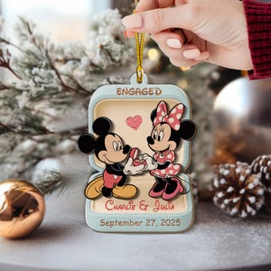May include: Wooden Christmas ornament featuring Mickey Mouse proposing to Minnie Mouse. The ornament is shaped like a ring box with the word "ENGAGED" at the top. The names "Charlie & Julia" and the date "September 27, 2025" are also included.