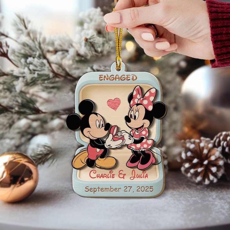 May include: Wooden Christmas ornament featuring Mickey Mouse proposing to Minnie Mouse. The ornament is shaped like a ring box with the word "ENGAGED" at the top. The names "Charlie & Julia" and the date "September 27, 2025" are also included.
