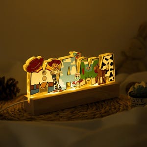 May include: Illuminated "MAMA" sign featuring characters from the Toy Story franchise. The letters are decorated with images of Woody, Jessie, and other characters. The sign is mounted on a wooden base and emits a warm, inviting glow.