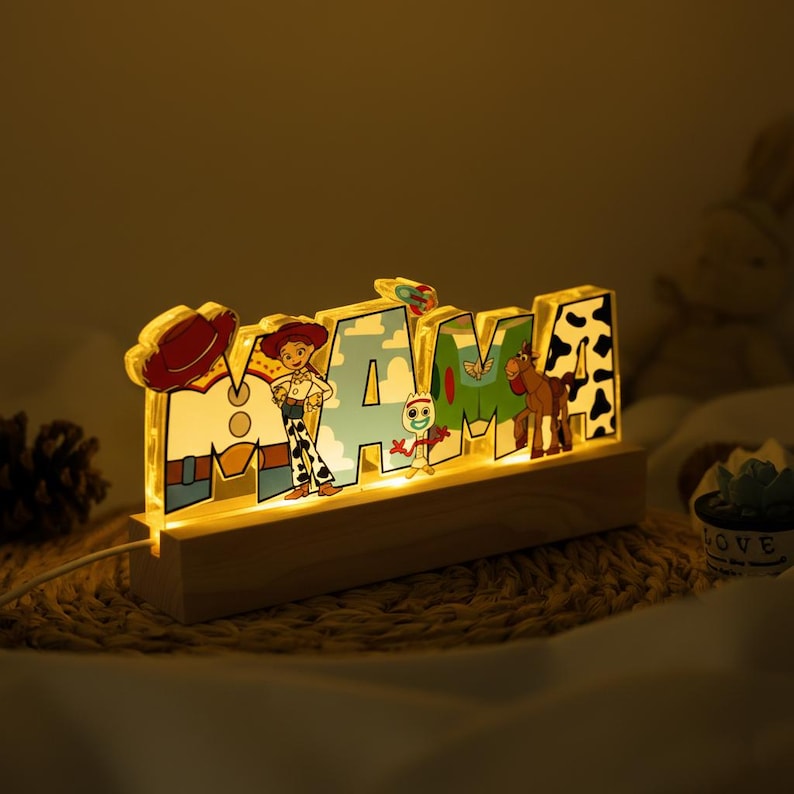 May include: Illuminated "MAMA" sign featuring characters from the Toy Story franchise. The letters are decorated with images of Woody, Jessie, and other characters. The sign is mounted on a wooden base and emits a warm, inviting glow.