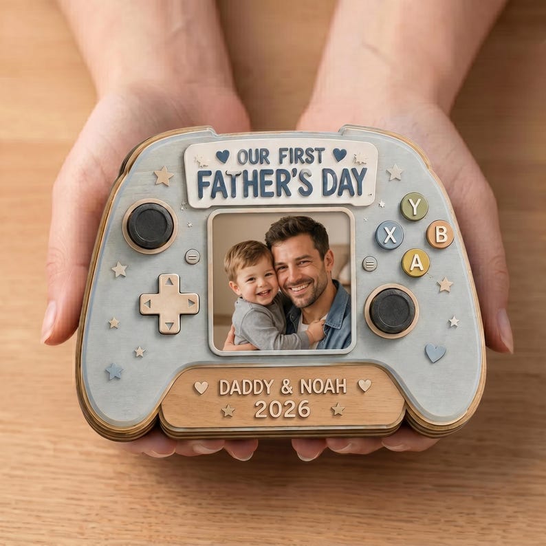 May include: A wooden Father's Day photo frame shaped like a video game controller. The frame features a photo of a father and child, with the text "OUR FIRST FATHER'S DAY" and "DADDY & NOAH 2026". The controller is light blue and wood-toned.