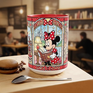 Puede incluir: Una taza de cer&aacute;mica blanca con interior rojo y un dise&ntilde;o de vidriera que representa a Minnie Mouse leyendo un libro. El dise&ntilde;o incluye un castillo, estrellas y un lazo rojo con lunares. Una cuchara y un pastelito est&aacute;n sobre la mesa de madera.