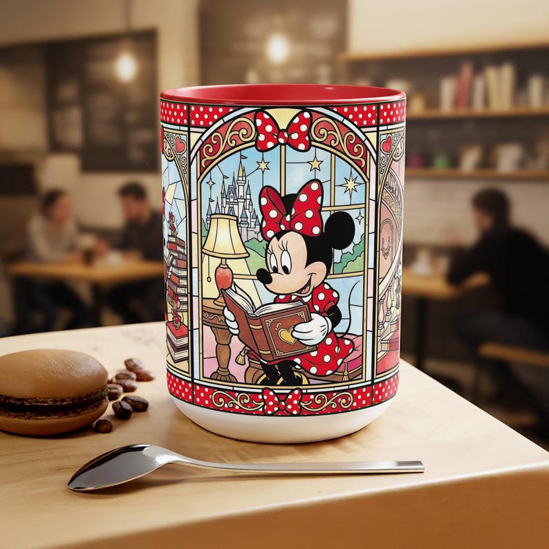 Puede incluir: Una taza de cer&aacute;mica blanca con interior rojo y un dise&ntilde;o de vidriera que representa a Minnie Mouse leyendo un libro. El dise&ntilde;o incluye un castillo, estrellas y un lazo rojo con lunares. Una cuchara y un pastelito est&aacute;n sobre la mesa de madera.