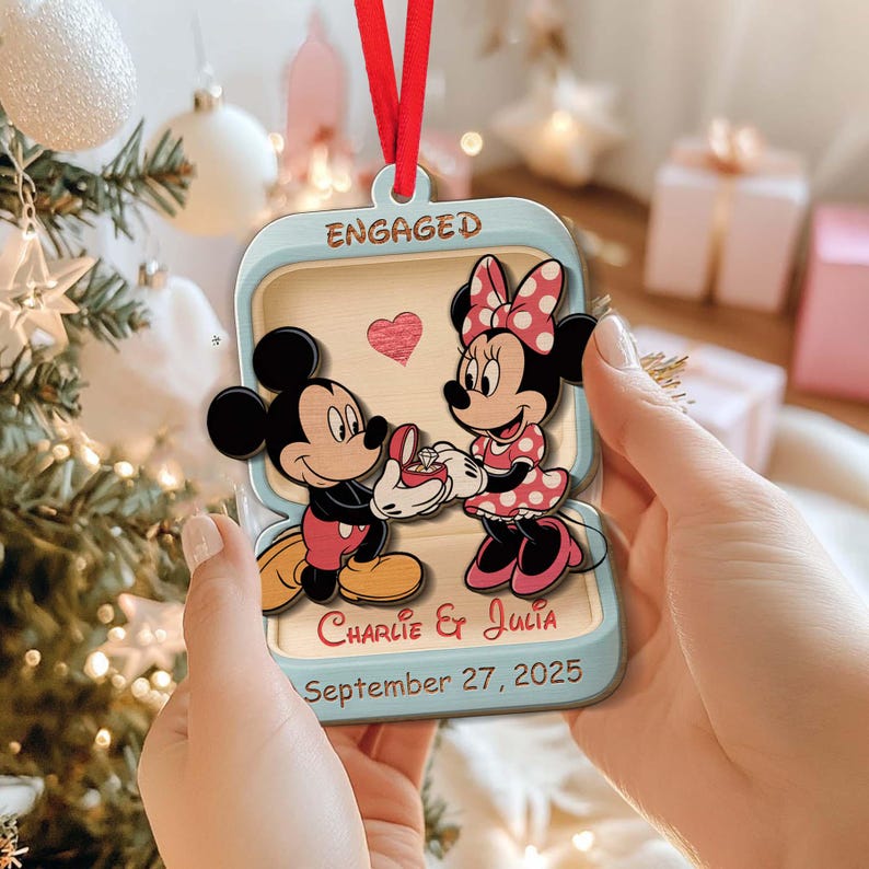 May include: A light blue and wood-toned ornament featuring Mickey and Minnie Mouse. Mickey Mouse is offering a ring to Minnie. The ornament reads "Engaged", "Charlie & Julia", and "September 27, 2025". A red ribbon is attached for hanging.