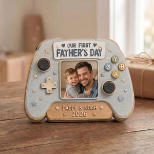 May include: A wooden photo frame shaped like a video game controller. The frame is light blue with decorative stars and hearts. It features a photo of a man and a child, with the text "OUR FIRST FATHER'S DAY" and "DADDY & NOAH 2026".