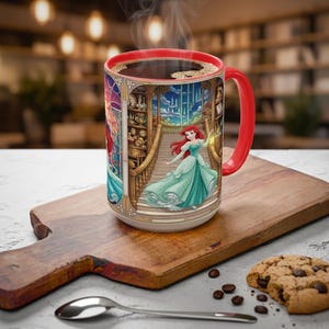 Pu&ograve; includere: Una tazza da caff&egrave; in ceramica con manico e bordo rossi, piena di caff&egrave;. La tazza presenta un'illustrazione colorata di una principessa in un abito verde acqua, con i capelli rossi, che scende una scala. La tazza &egrave; su un tagliere di legno.