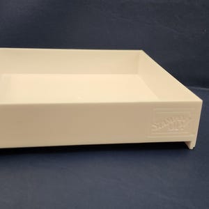 Puede incluir: Una bandeja rectangular blanca con un perfil bajo, con un borde ligeramente elevado y una base plana. La bandeja tiene un diseño minimalista y probablemente está hecha de plástico. El nombre de la marca está grabado en relieve en el lateral.