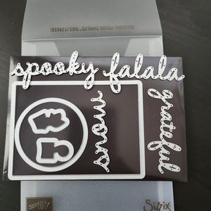 May include: A set of white metal die cuts for scrapbooking and card making. The dies include the words "spooky", "falala", "grateful", and "snow".