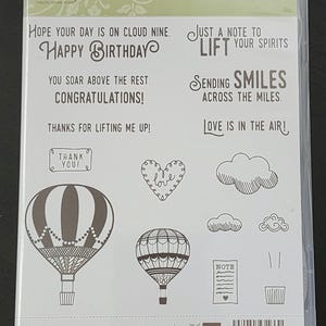 Puede incluir: Un conjunto de 16 sellos de goma transparente con un tema de mensajes alentadores y globos aerostáticos. Los sellos incluyen frases como "Espero que tu día esté en la nube. Feliz cumpleaños", "Solo una nota para levantar tu ánimo. Enviando sonrisas a través de las millas. ¡El amor está en el aire!" y "¡Gracias por levantarme!"