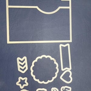 Peut inclure: Formes blanches découpées sur une surface bleue. Comprend un grand rectangle avec une section incurvée, une bannière, un cœur, une étoile, un cercle festonné et des découpes de mots lisant "thx", "xoxo" et "yay".