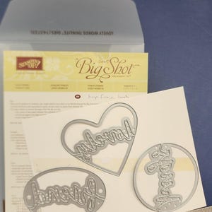 May include: Silver metal word dies in heart, oval, and circle shapes, featuring the words "Loved" and "Forever". A stainless steel ruler measures the dies in centimetres. The background includes a Big Shot die-cutting machine package.