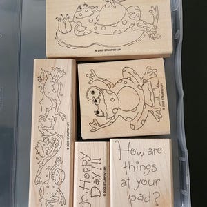 Peut inclure: Ensemble de cinq tampons en caoutchouc en bois représentant des grenouilles et le texte "Oh Hoppy Day!!!" et "How are things at your pad?"