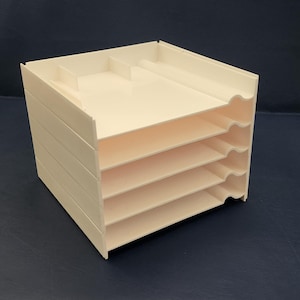 Puede incluir: Un organizador de escritorio crema de varios niveles con cinco ranuras horizontales para papeles y documentos. El nivel superior tiene un área empotrada. El organizador está hecho de un material opaco sólido y se encuentra sobre una superficie azul oscuro.