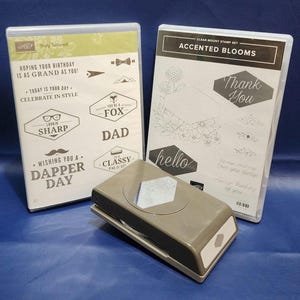 May include: A collection of crafting supplies, including two clear mount stamp sets and a gray paper punch. One stamp set reads "Truly Tailored" with phrases like "Dapper Day" and "Lookin' Sharp." The other set says "Accented Blooms."