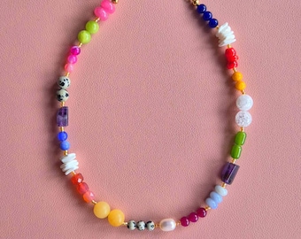 May include: A colorful beaded necklace with a gold chain. The necklace features a variety of beads in shades of pink, green, blue, white, red, yellow, and purple. A pearl is also included in the design. The necklace is displayed on a pink background.