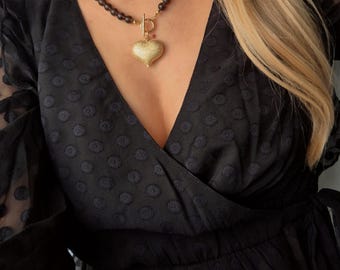 Op de afbeelding: Een gouden hartvormige hangerketting met een zwarte kralenketting. De hartvormige hanger heeft een textuur en is bevestigd aan een gouden sluiting. De ketting wordt gedragen over een zwarte wikkel-top.