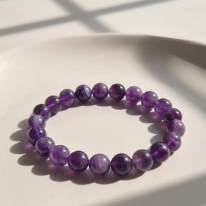 Puede incluir: Un brazalete de cuentas redondas de amatista pulida. Las cuentas son de un color púrpura intenso. El brazalete está sobre un plato redondo de color claro. El brazalete es elástico y tiene una circunferencia de unos 18 cm.