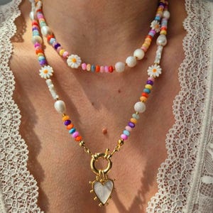 Può includere: Collana a due strati con perline colorate, perle bianche e accenti a margherita. La fila inferiore presenta un ciondolo a cuore dorato con un centro bianco. La collana è indossata su uno sfondo di pizzo bianco.