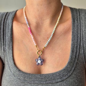 Op de afbeelding: Een ketting met een gouden sluiting en een paarse bloemhanger. De ketting heeft een verloop van pastelkleuren, waaronder roze, blauw en geel. De bloemhanger heeft een wit midden.