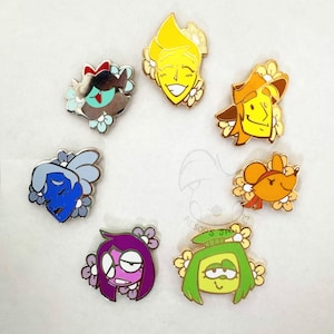 Puede incluir: Una colección de siete pines de esmalte coloridos dispuestos en círculo, cada uno con una cabeza de personaje de dibujos animados única con detalles florales. Los personajes están representados en tonos vibrantes de amarillo, naranja, verde, morado, azul y marrón.
