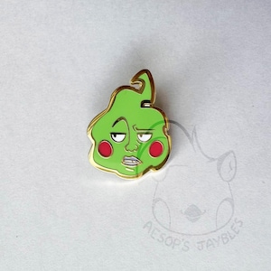 Op de afbeelding: Een groene emaille pin met een gouden rand, die een stripfiguur afbeeldt met een humeurige uitdrukking en rode wangen. De pin heeft een wervelvormige bovenkant en staat tegen een witte achtergrond.