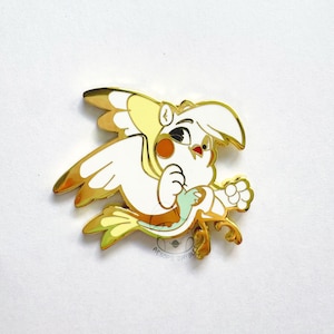 Op de afbeelding: Een goudkleurige emaille pin met een cartoonvogel met witte, gele en bruine kleuren. De vogel heeft een verbaasde uitdrukking en houdt een klein, lichtgroen object vast. De pin heeft een sierlijk ontwerp aan de onderkant en de tekst "AESOP'S SHYBUS".