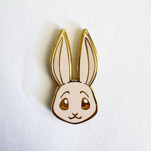 May include: A gold-toned metal enamel pin shaped like a rabbit head. The rabbit has light pink enamel fill, large brown eyes with white highlights, and long upright ears. The metal edges are polished gold, outlining the ears, face, and features.
