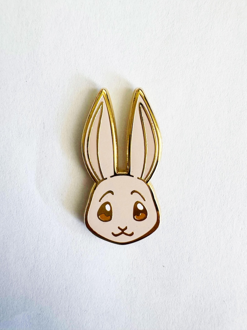 May include: A gold-toned metal enamel pin shaped like a rabbit head. The rabbit has light pink enamel fill, large brown eyes with white highlights, and long upright ears. The metal edges are polished gold, outlining the ears, face, and features.