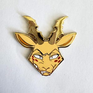 May include: A gold-toned metal enamel pin shaped like the head of a horned creature. The design features a pale yellow face with pointed ears, dark red spots on the cheeks, sharp white teeth, and metallic gold horns with a textured edge.