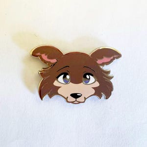 May include: A metal enamel pin shaped like the head of a brown wolf with large blue eyes, pink inner ears, and a light tan muzzle. The design features a dark brown nose and a silver-colored metal outline that defines the edges of the character.