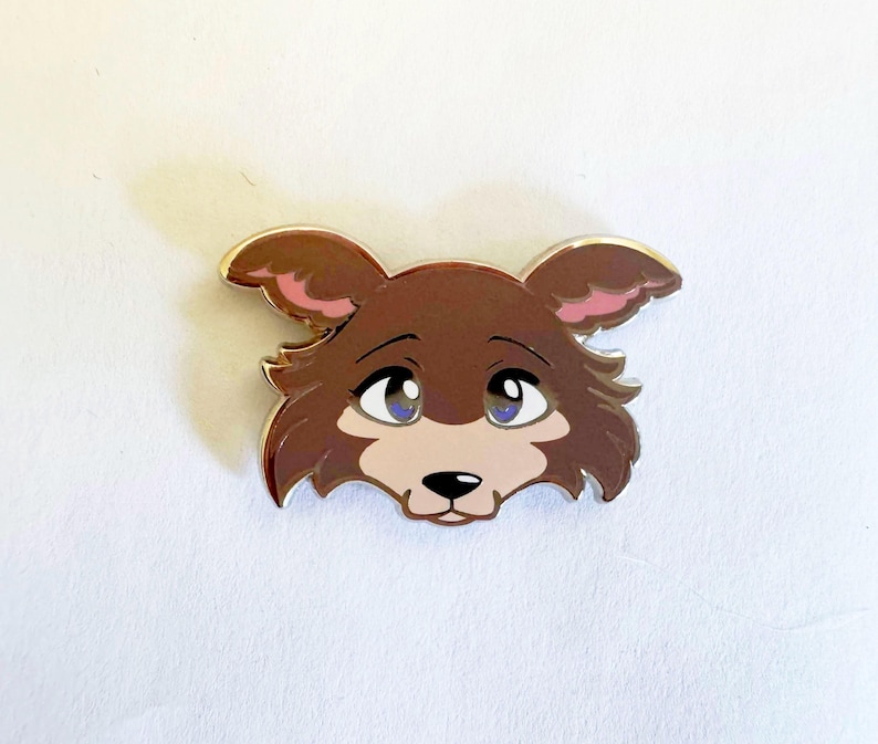 May include: A metal enamel pin shaped like the head of a brown wolf with large blue eyes, pink inner ears, and a light tan muzzle. The design features a dark brown nose and a silver-colored metal outline that defines the edges of the character.